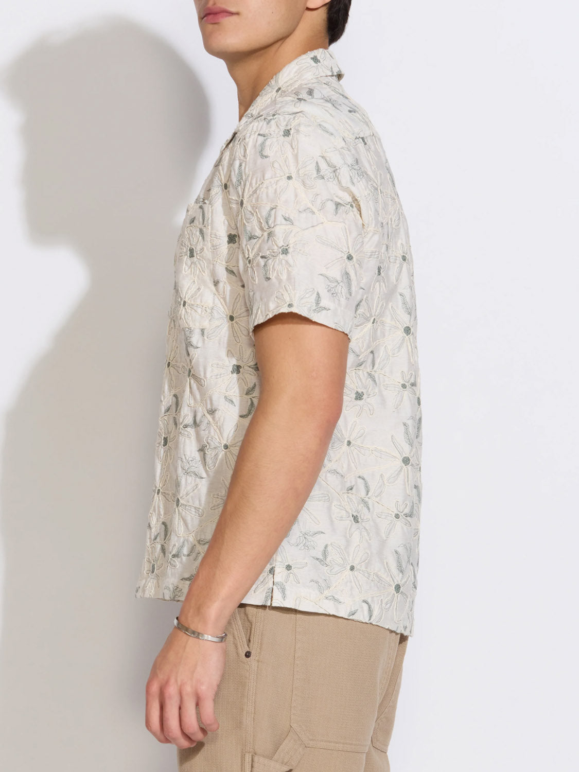 A person in the Civil Society Zanzibar Embroidered Appliqué Resort Shirt and beige pants stands sideways against a white background. The shirt's Cuban collar and floral appliqué offer a relaxed vibe; their face is partially out of frame.