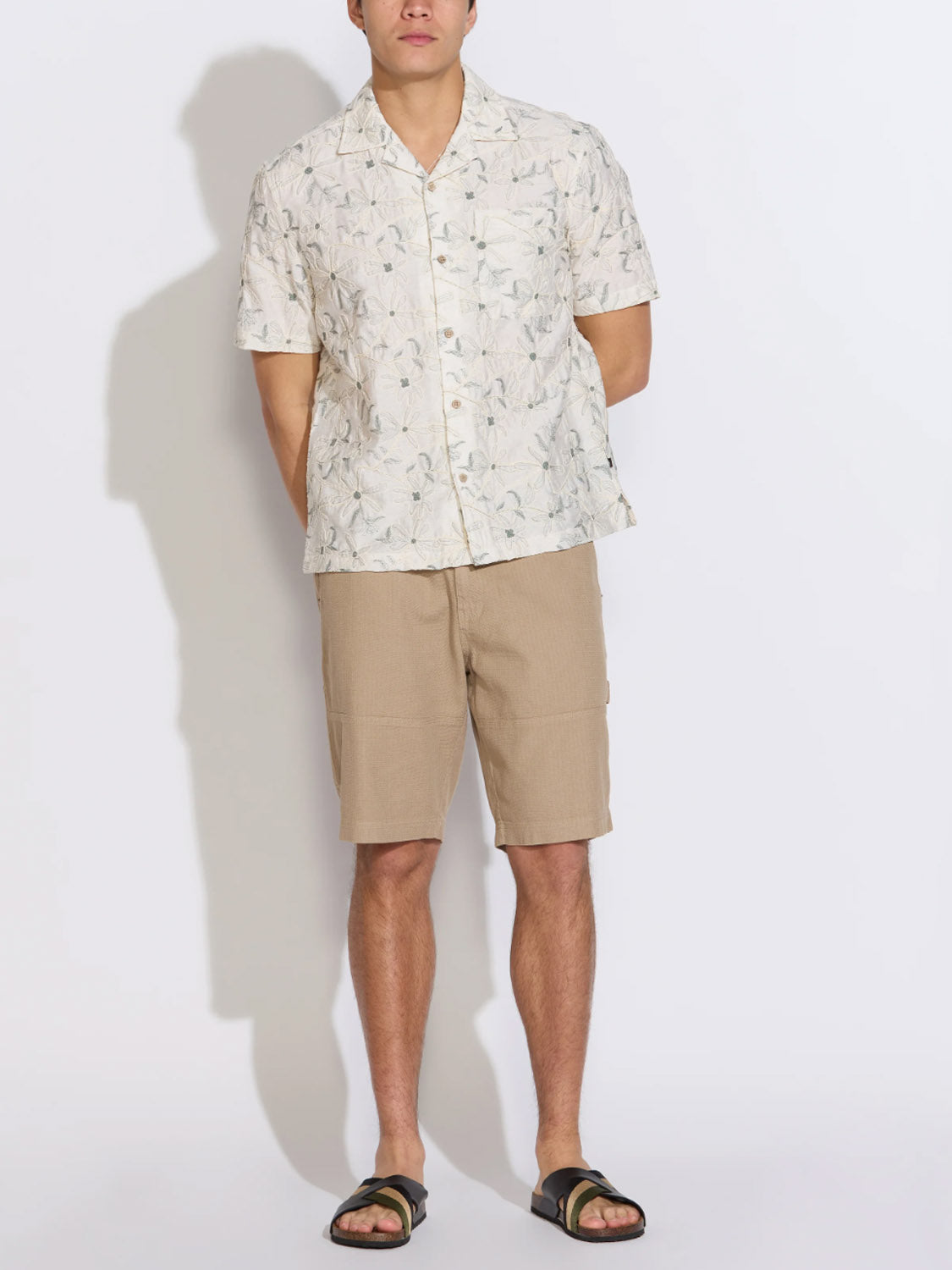 A person faces forward against a plain white background, wearing the Civil Society Zanzibar Embroidered Appliqué Resort Shirt, styled with tan knee-length shorts and black sandals.