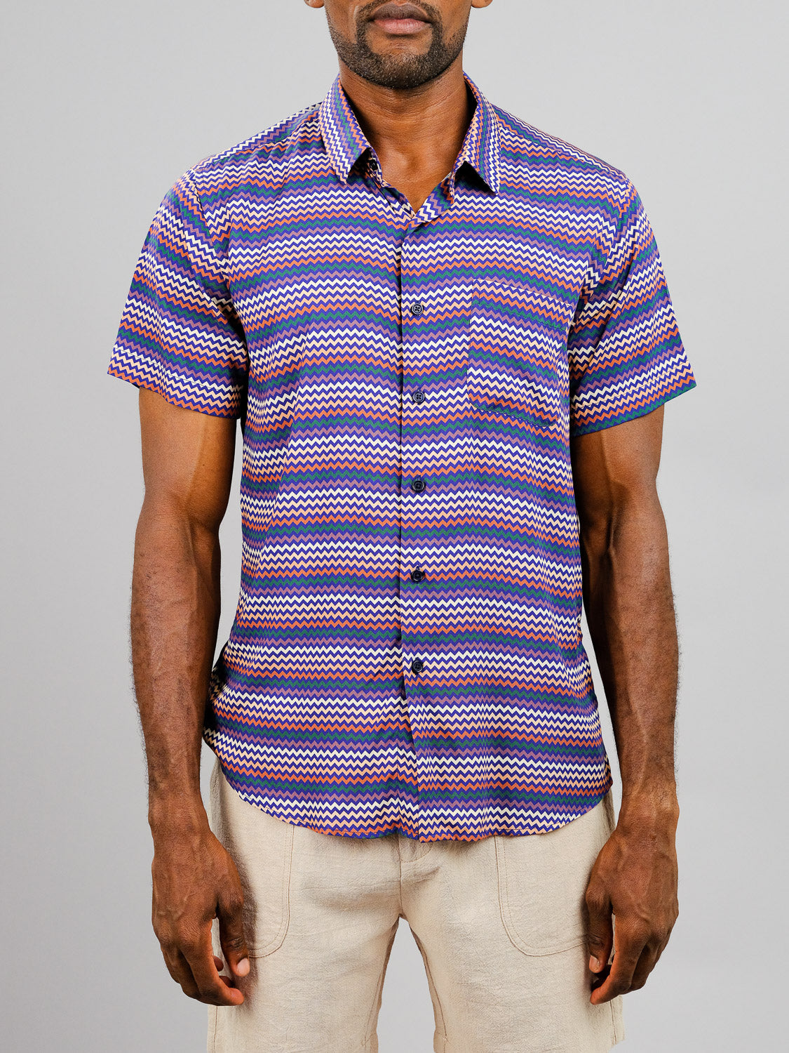 A man models the Ziggy Printed Rayon Shirt by ID Menswear, featuring vibrant purple, blue, and pink zigzag stripes and short sleeves, paired with beige pants against a plain light gray background. His face is partially out of view.