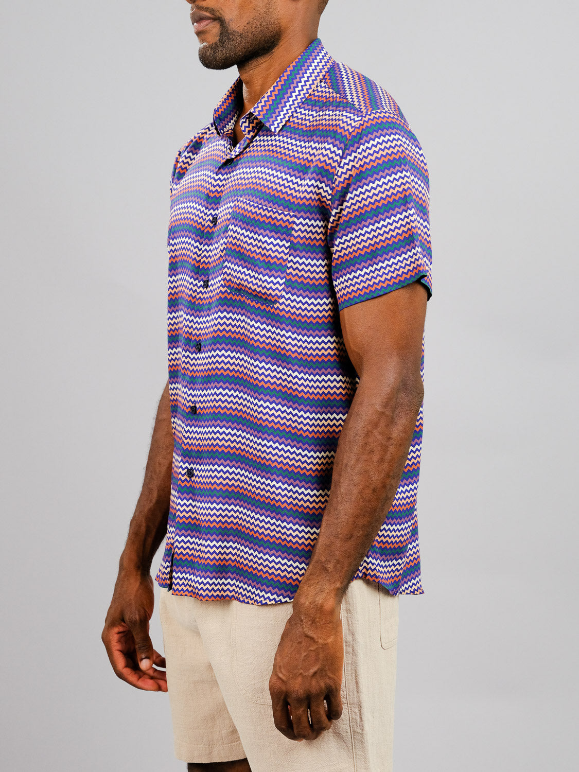 A man wears the ID Menswear Ziggy Printed Rayon Shirt—lightweight with blue, red, and white zigzag stripes—and light beige shorts, standing against a plain gray background.