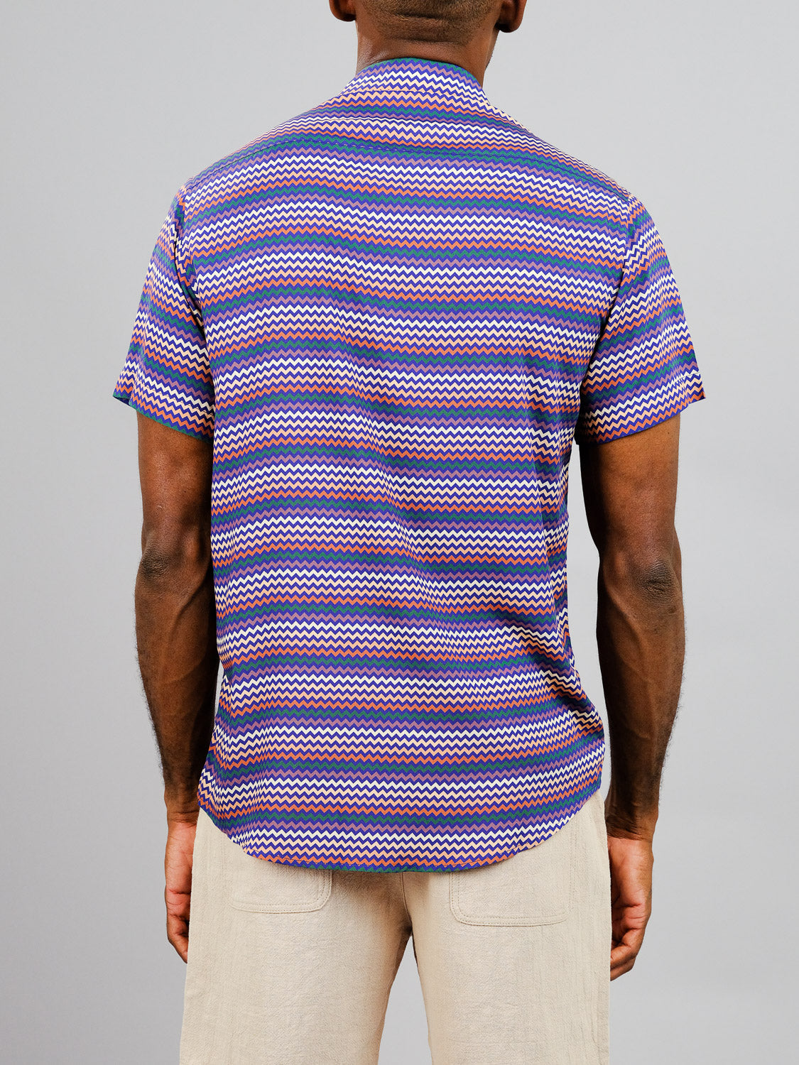 A person is shown from the back wearing the ID Menswear Ziggy Printed Rayon Shirt, featuring a vibrant purple, pink, and blue zigzag print, paired with light beige pants against a plain light gray background.