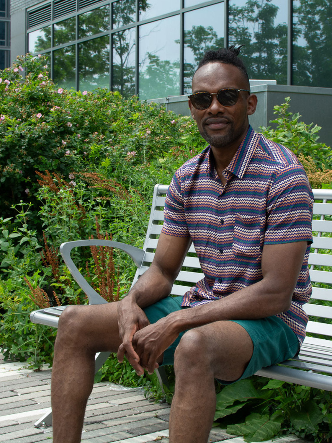 Wearing the Ziggy Printed Rayon Shirt by ID Menswear and green shorts, a man in sunglasses sits on an outdoor bench near bushes and flowers, with a building behind him—showcasing the perfect warm-weather style.