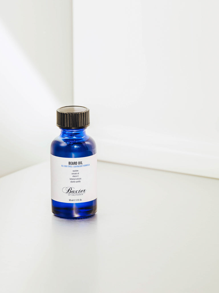 A blue glass bottle of Baxter Beard Oil with a black cap rests on a light surface against a white background, softly lit by natural light.