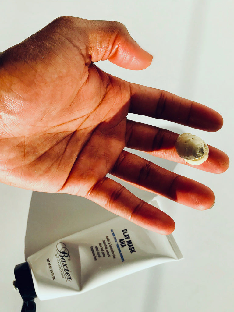 A hand displays a small amount of Baxter Clay Mask AHA on the palm, with the white tube partially visible underneath. The scene is brightly lit, casting subtle shadows.