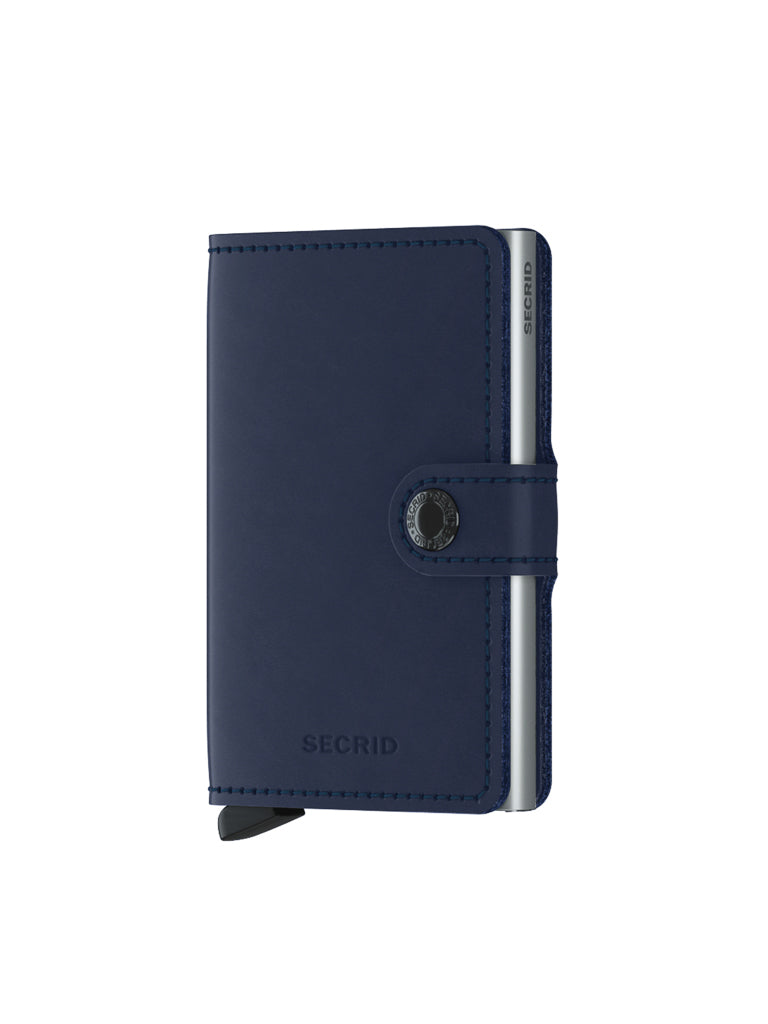 The Secrid Miniwallet Original by Secrid, in navy blue with a button snap closure and visible aluminum card protector on the right side, stands upright on a white background, offering RFID security in a compact design.