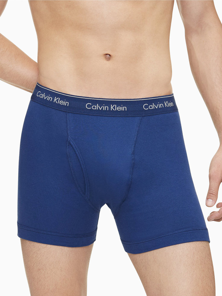 A person models Calvin Klein NB4003 3-Pack Cotton Classic Boxer Brief featuring a black waistband with white branding. The blue boxer brief covers the torso and upper legs, shown against a plain background.