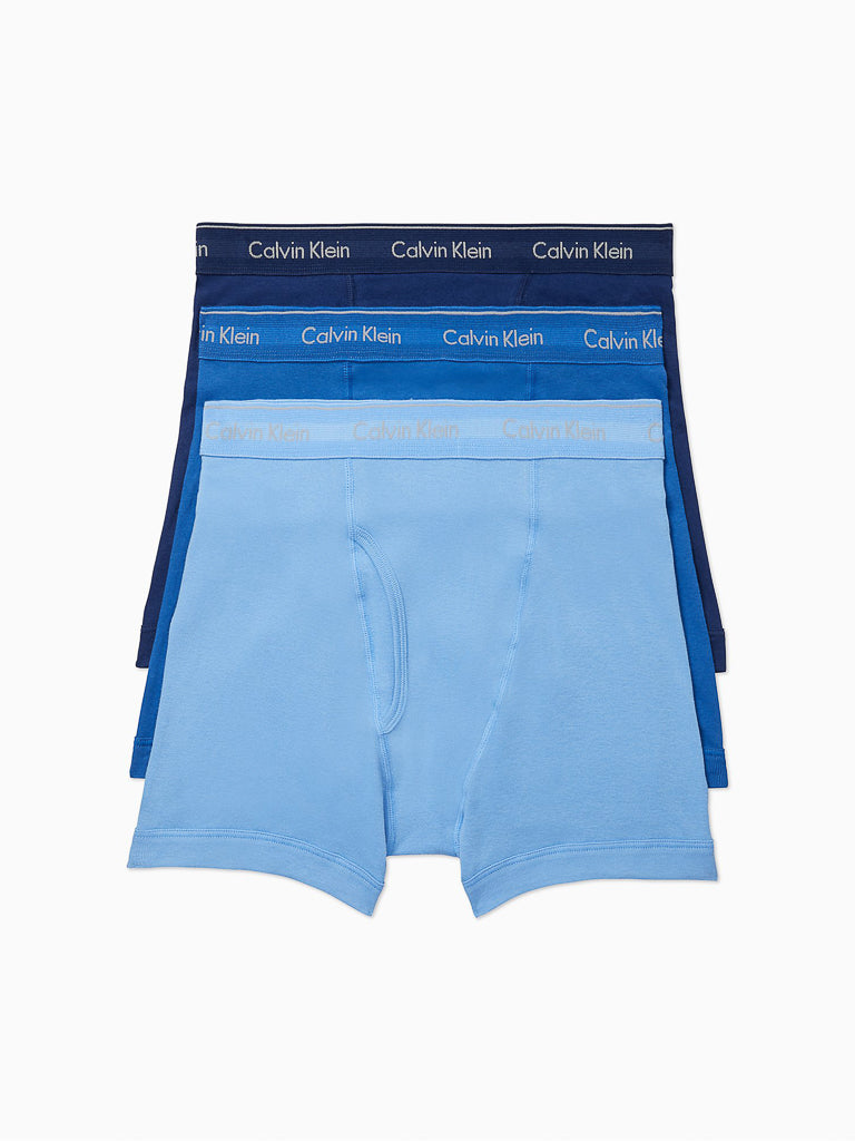 A trio of Calvin Klein NB4003 Cotton Classic Boxer Briefs is stacked to highlight the signature logo waistbands, featuring pure cotton fabric in a gradient of blue shades from light to dark.