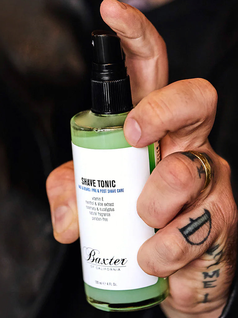 A tattooed hand with a gold ring holds a green bottle of Baxter Shave Tonic, a cooling aftershave with a black spray nozzle and white label, designed to help prevent ingrown hairs.