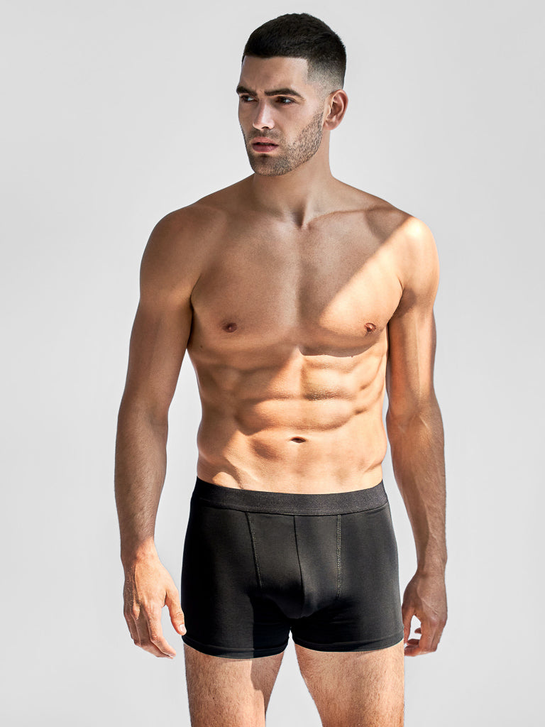 A muscular man with short dark hair stands against a plain background, wearing black Bread & Boxer briefs from the Bread and Boxers 3 Pack. Made from organic cotton, they have an elastic waistband. He gazes to his left as shadows highlight his physique.