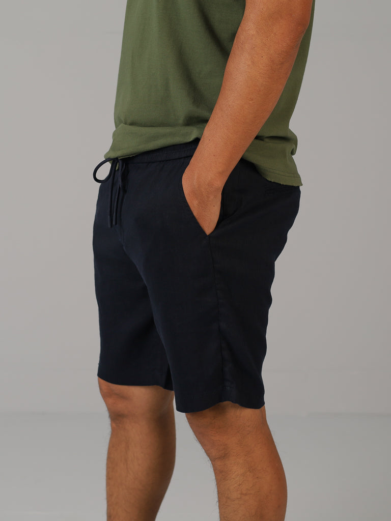 A person in a green t-shirt and black shorts with their right hand in a pocket showcases ID Menswears Driggs Linen Blend Short. These shorts, ideal for warm weather with their casual style and drawstring waist, are set against a gray background.