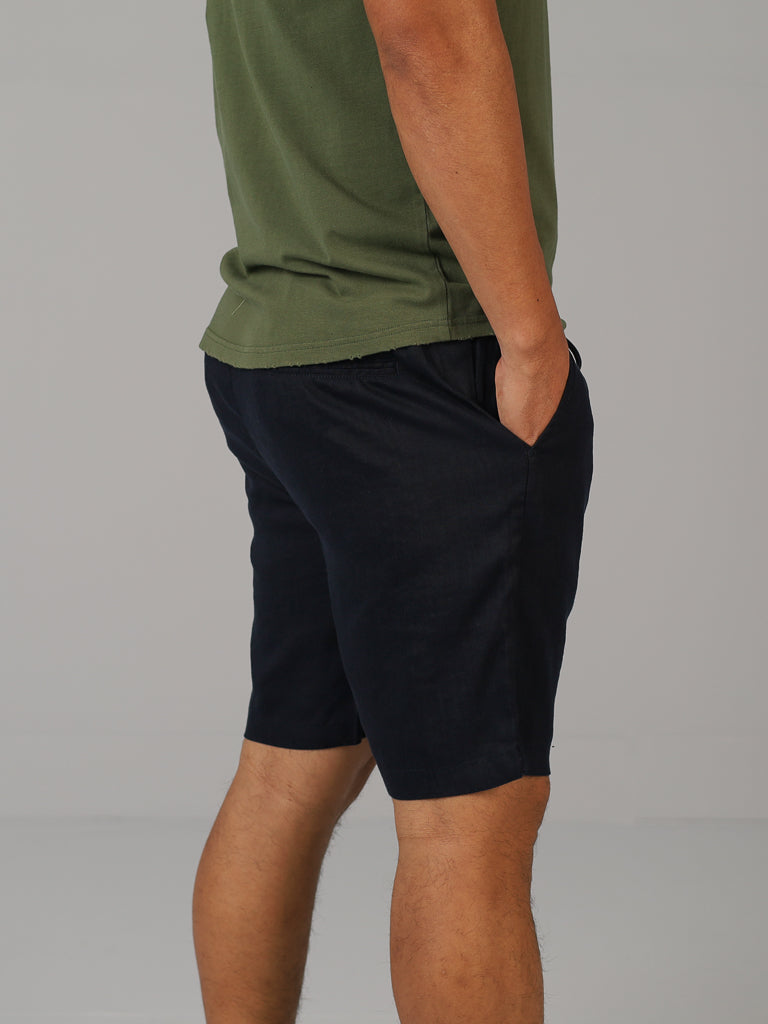 A person is standing sideways in a green T-shirt and ID Menswears Driggs Linen Blend Shorts in black, ideal for warm weather. Their left hand casually rests in the pocket against a plain white background.