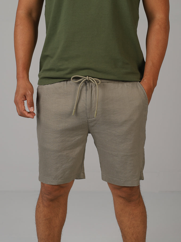 A person wearing an olive green shirt and ID Menswears light gray Driggs Linen Blend Short stands with hands in pockets against a plain gray background, perfectly embodying the essence of warm-weather clothing.