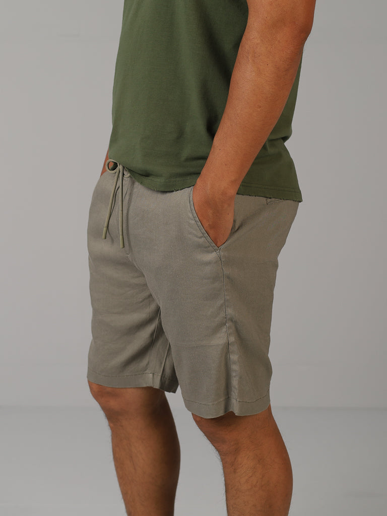 A person is dressed in a green T-shirt and the Driggs Linen Blend Short by ID Menswear. Their hands are tucked in pockets against a plain light gray background. This casual, comfortable outfit is perfect for warm weather and is shown from shoulders down with no visible head.