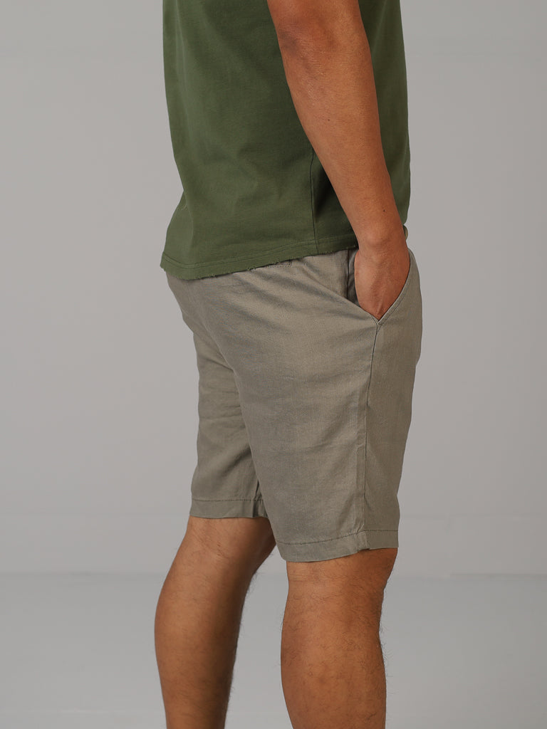A person in a green shirt and ID Menswears Driggs Linen Blend Short stands with one hand in their pocket, facing sideways against a plain gray backdrop, embodying a relaxed vibe ideal for warm weather.