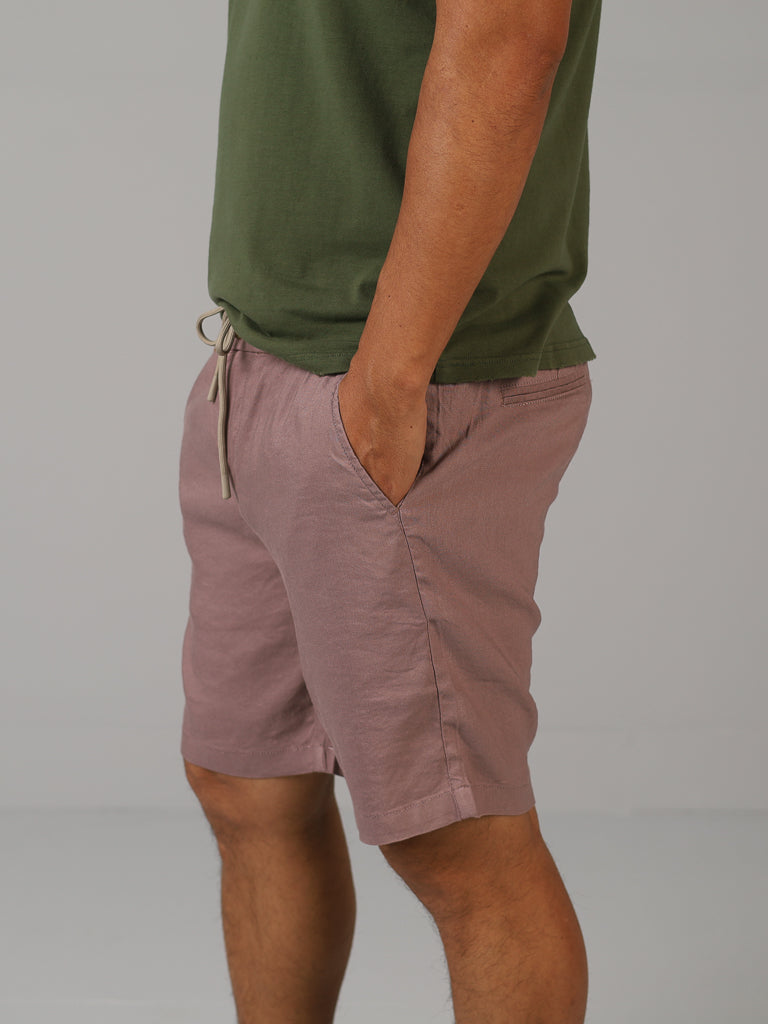 A person in a green t-shirt and ID Menswears Driggs Linen Blend Shorts—the mauve shorts, perfect for warm weather, feature hands-in-pockets and a visible drawstring—stands against a plain gray background.