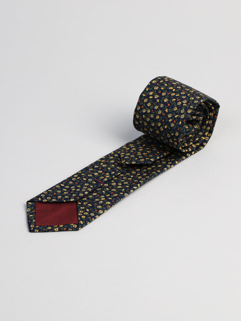 The Japanese Printed Hana NYGR Tie by ID Menswear is handmade in Brooklyn from 100% cotton, featuring a dark floral print with yellow and red accents, a solid burgundy back patch, and is displayed on a light gray surface.