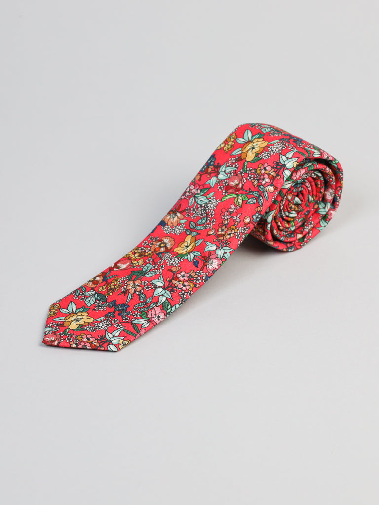 The P. Bouquet Cotton Tie by ID Menswear features a bright red background with a colorful floral pattern, handmade in Brooklyn. Perfect for business formal looks, its displayed rolled on a plain white surface.