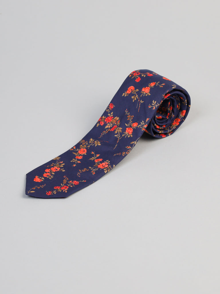 The Liberty of London Tana Lawn™ Elizabeth Tie by ID Menswear is handmade in Brooklyn from soft navy blue Tana Lawn cotton with a red and green floral pattern, shown rolled on a light gray surface.