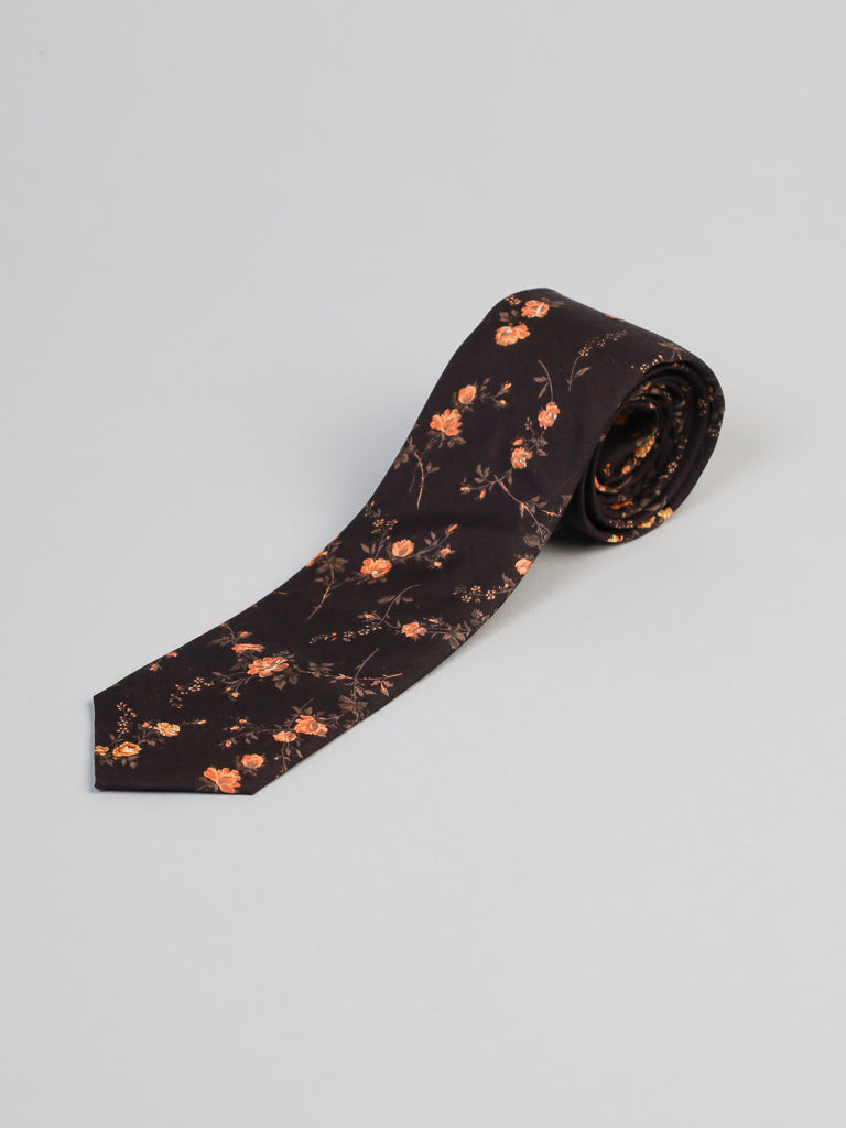 A Liberty of London Tana Lawn™ Elizabeth Tie by ID Menswear, featuring a dark brown base and orange floral pattern, handmade in Brooklyn from fine Tana Lawn cotton, is rolled at one end and displayed on a light gray background.
