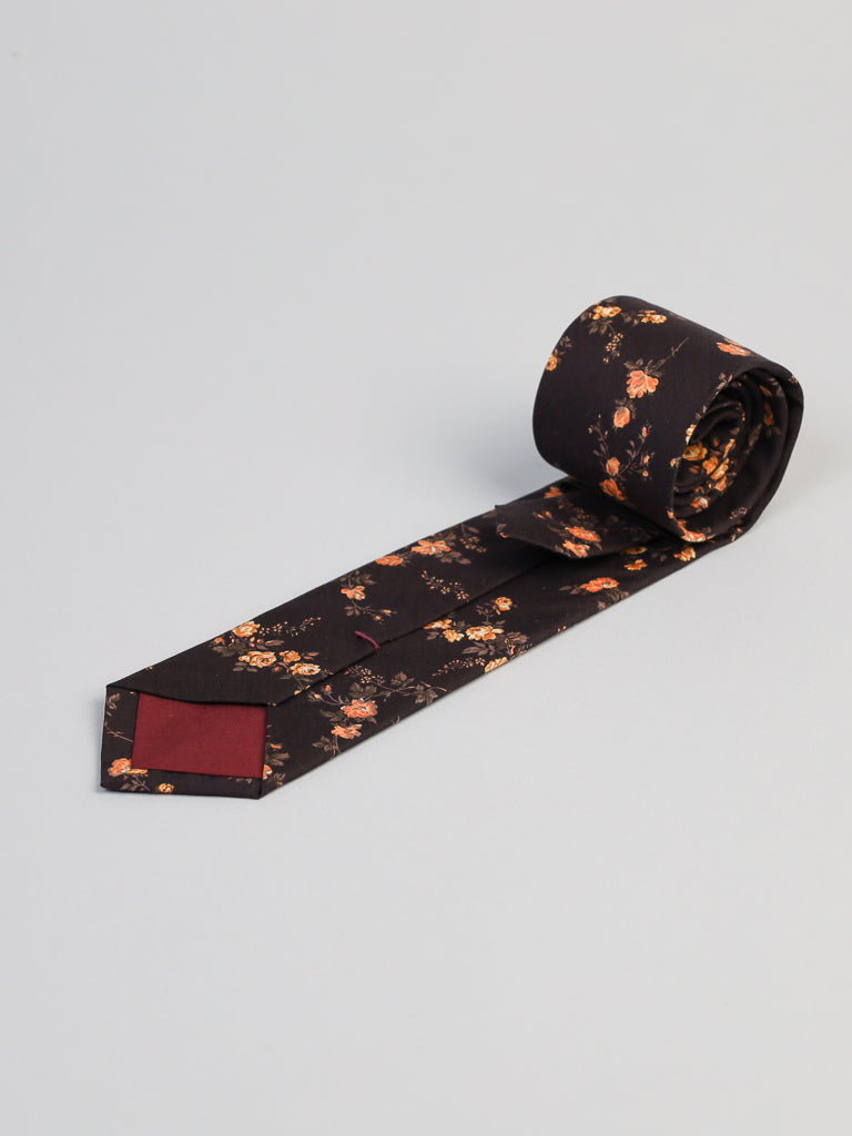 The ID Menswear Liberty of London Tana Lawn™ Elizabeth Tie features a dark floral pattern with orange and yellow flowers, a red accent at the tip, and is handmade in Brooklyn, shown rolled on a light gray background.