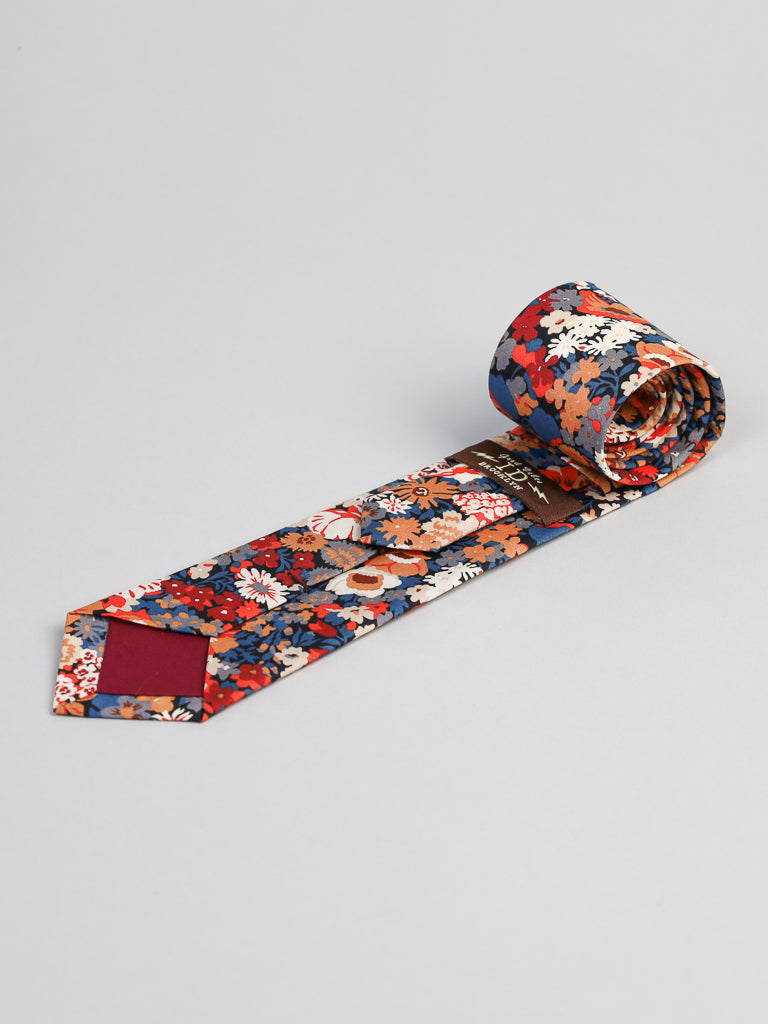 The Liberty of London Tana Lawn™ Kanazawa Tie by ID Menswear, handmade in Brooklyn, features a vibrant red, blue, white, and orange floral pattern on a dark background and is shown rolled up on a light gray surface.