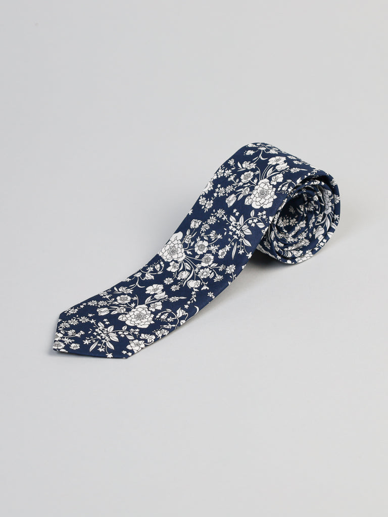 The Liberty of London Tana Lawn™ Navy Lawn Tie by ID Menswear, featuring a white floral pattern and silk-like touch, is rolled at one end and displayed on a plain light gray background.