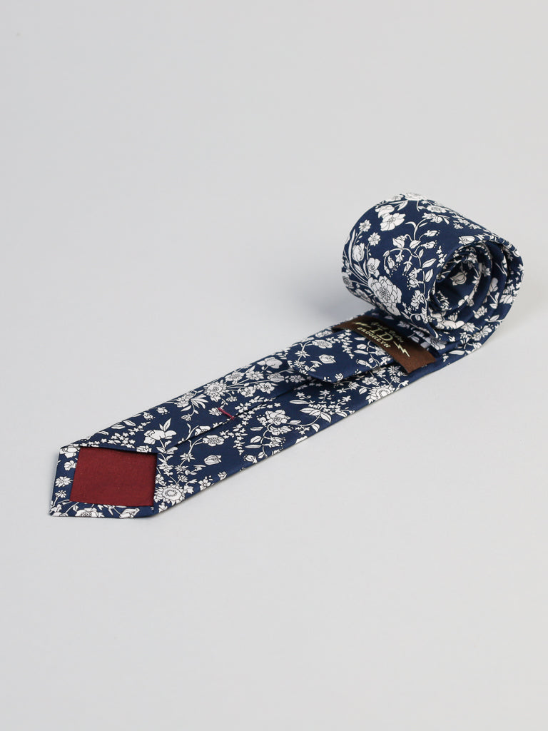 The Liberty of London Tana Lawn™ Navy Lawn Tie by ID Menswear is a handmade rolled navy tie with a white floral pattern and maroon inner tip, shown against a light gray background.