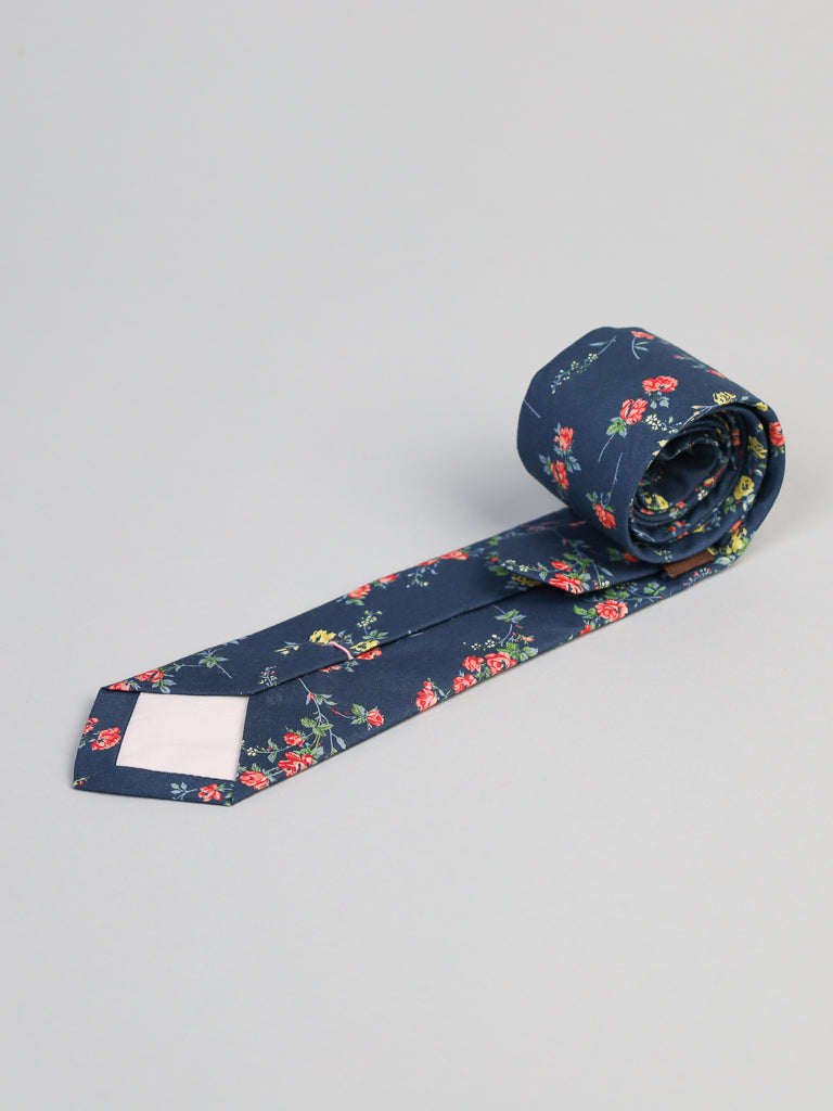 The Liberty of London Tana Lawn™ Elizabeth Tie by ID Menswear—navy blue, handmade in Brooklyn with a red, yellow, and green floral pattern—comes neatly rolled on a light gray background.