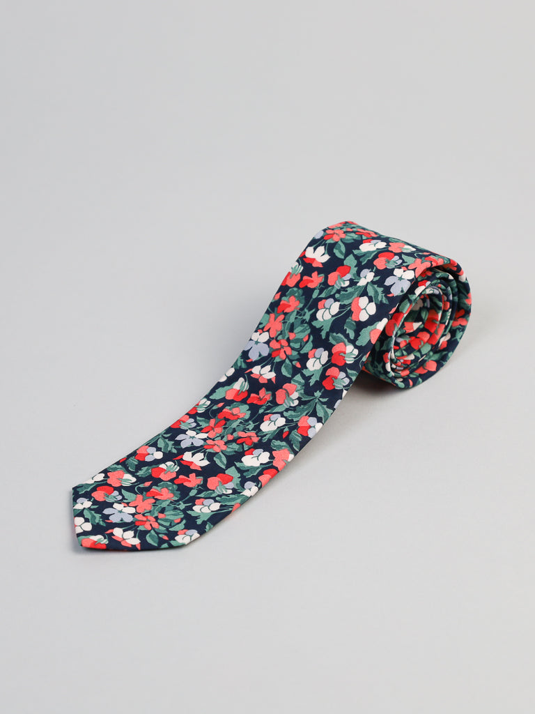 The Liberty of London Tana Lawn™ Classic Tie by ID Menswear features a colorful red, white, and green floral print on dark cotton—handmade in Brooklyn and shown rolled up on a light gray surface.