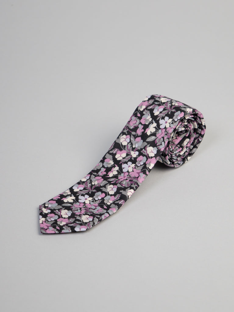 The ID Menswear Liberty of London Tana Lawn™ Sarah P. Tie features a floral pattern in pink, white, and lavender on black, crafted from fine cotton and rolled neatly on a plain light gray surface.