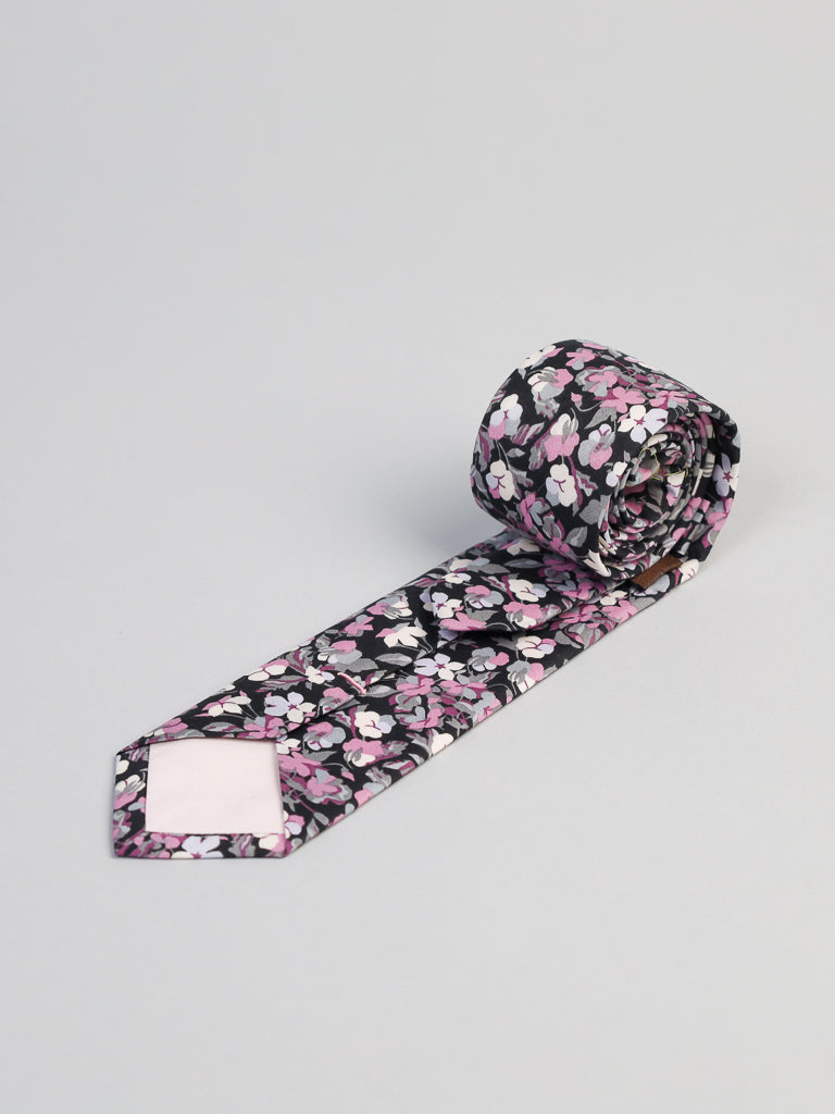 The ID Menswear Liberty of London Tana Lawn™ Sarah P. Tie features a black background with pink, white, and purple floral patterns in luxurious cotton, shown rolled up on a light gray surface.