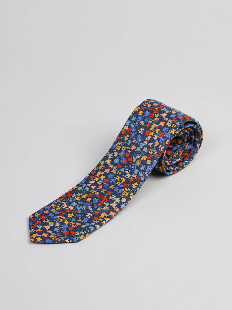 A Liberty of London Tana Lawn™ Gizmo Tie by ID Menswear, handmade in Brooklyn, features a dark blue background with a vibrant floral print in red, yellow, orange, and blue. The tie is rolled up and displayed on a plain light surface.