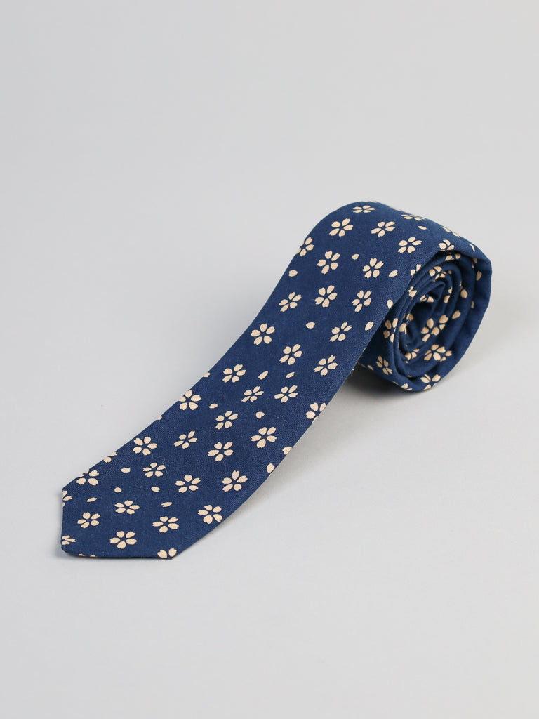The Japanese Printed Sakura Tie by ID Menswear is a navy blue 100% cotton tie with light floral designs, handmade in Brooklyn. Its shown rolled up on a plain light gray surface.