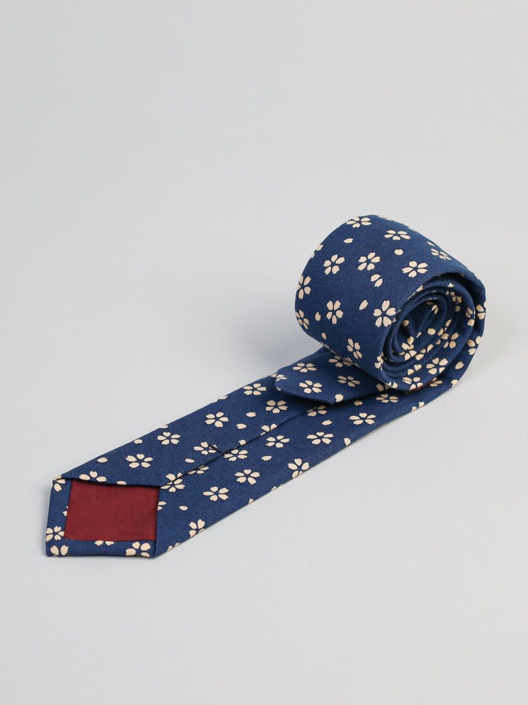 The Japanese Printed Sakura Tie by ID Menswear is a navy blue cotton tie with white floral print, handmade in Brooklyn, featuring red lining at the back tip. Shown rolled up on a light gray background.