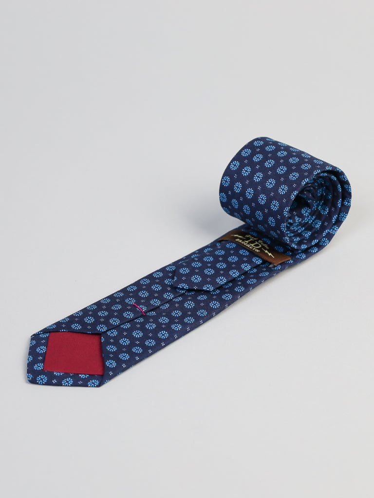 The Play Day Cotton Tie by ID Menswear is a navy blue tie with small light blue flowers, a red fabric patch under the tip, and a partially rolled design. Handmade in Brooklyn, its ideal for business formal wear and shown on a light gray background.