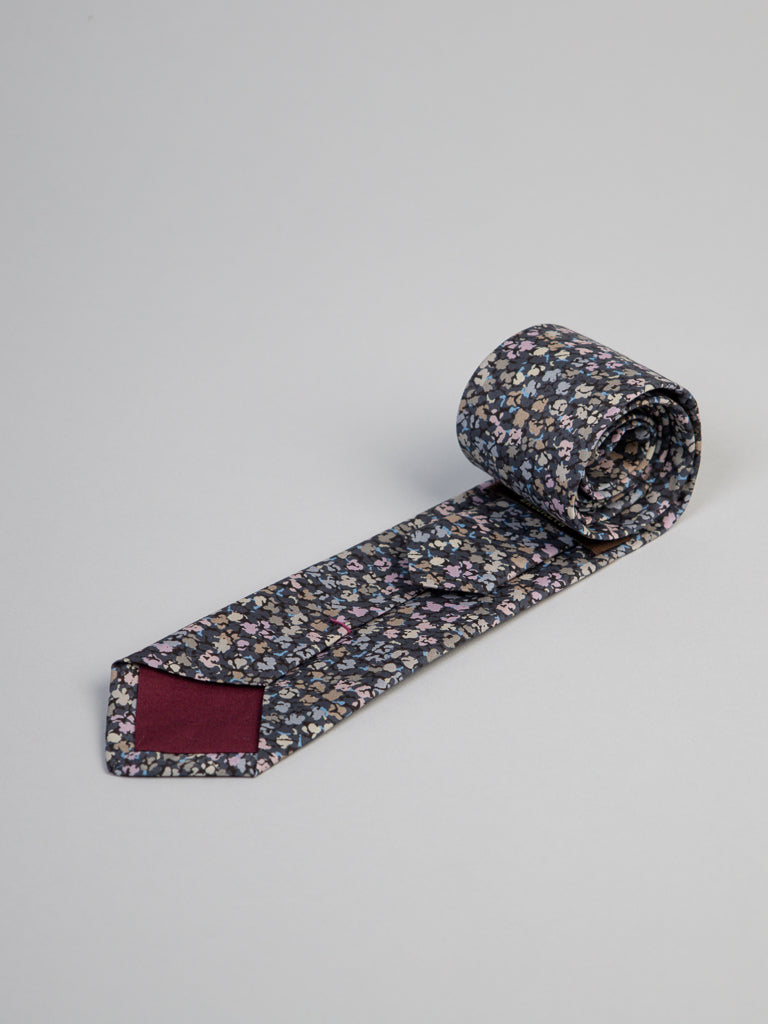 The ID Menswear Liberty of London Tana Lawn™ Gizmo Tie features a dark floral pattern with multicolored flowers and a burgundy tip, shown rolled up on a light gray background.