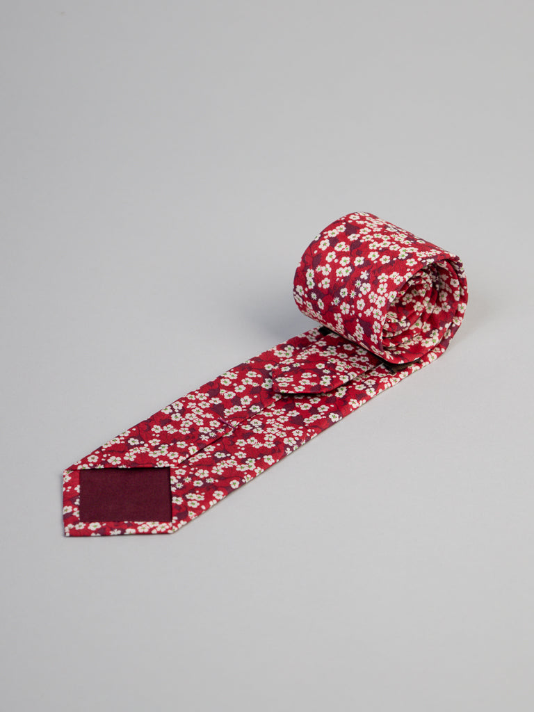 A rolled-up Liberty of London Tana Lawn™ Valeria Tie by ID Menswear, handmade in Brooklyn from cotton, featuring a small white floral pattern on red, displayed on a plain light gray background.