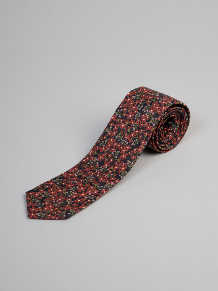 The ID Menswear Liberty of London Tana Lawn™ Anise Tie, featuring a red and black floral pattern and handmade in Brooklyn from luxurious cotton, is shown rolled up against a plain light gray background.