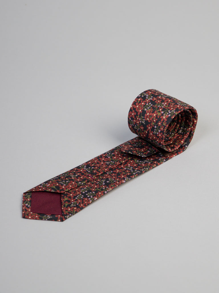 The Liberty of London Tana Lawn™ Anise Tie by ID Menswear features a dark red backdrop with a small multicolored floral print, is handmade in Brooklyn, and is shown rolled up on a light gray surface.
