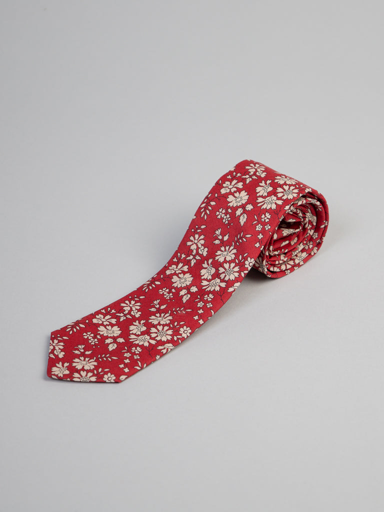 A red Liberty of London Tana Lawn™ Carpel Tie by ID Menswear, handmade in Brooklyn with a white floral pattern, is rolled up on a plain light gray background.