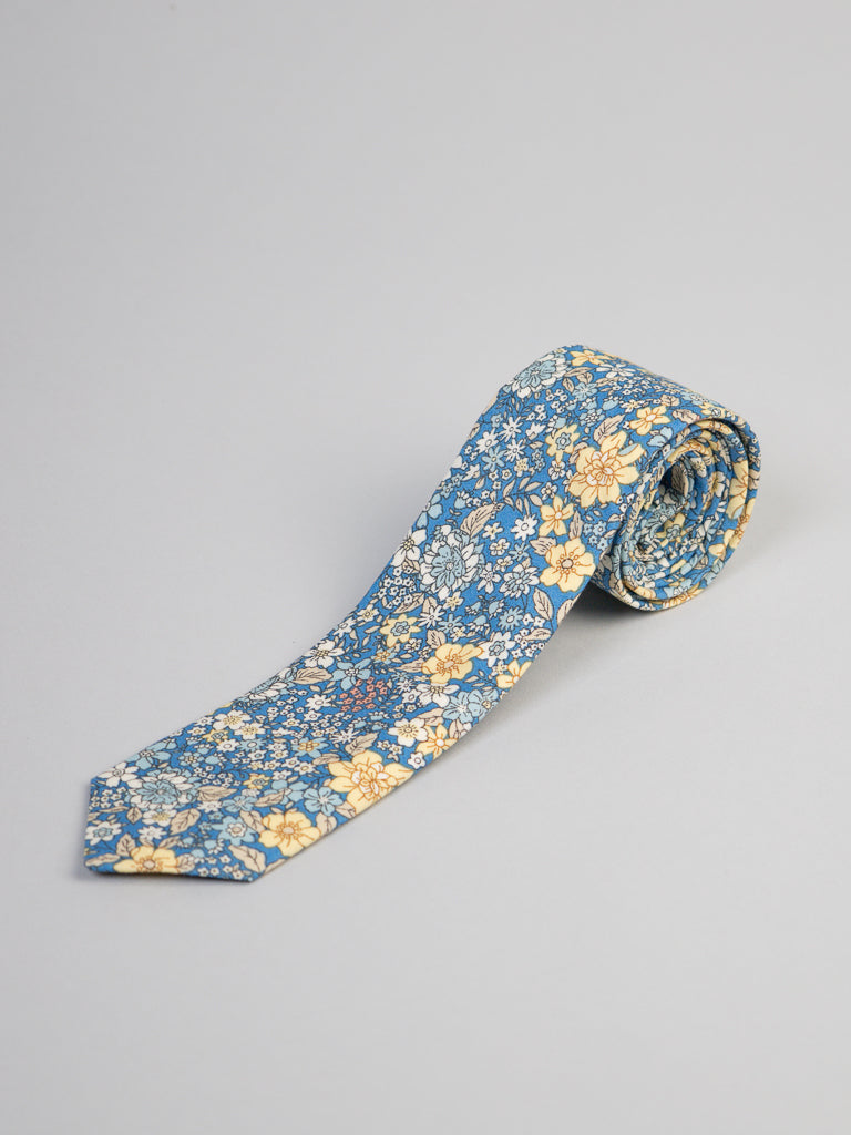 The ID Menswear Poplin Cotton Tie, handmade in Brooklyn with a blue background and small yellow and white floral patterns, is displayed on a plain light gray surface—perfect for business formal accessories.
