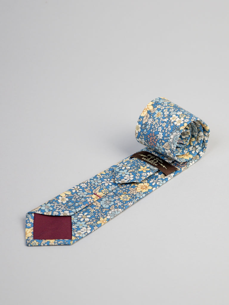The Poplin Cotton Tie by ID Menswear is a handmade cotton tie from Brooklyn, featuring a blue floral print with yellow, white, and light blue flowers—an ideal business formal accessory, shown on a plain light gray background.