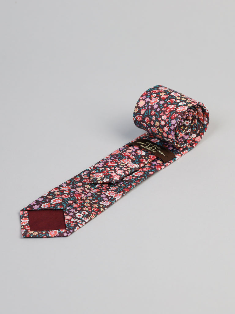 A rolled-up Liberty of London Tana Lawn™ Jo Tie by ID Menswear, featuring a colorful floral pattern on a dark background and crafted from fine cotton, is displayed on a light gray surface.