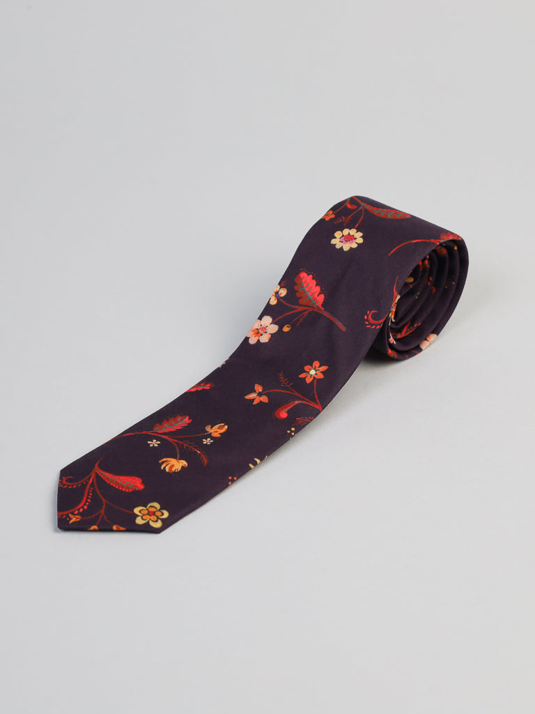 The Liberty of London Tana Lawn™ Swedish Tie by ID Menswear is a rolled-up, handmade dark purple tie with orange, red, and yellow floral patterns on luxurious cotton, displayed on a light gray background.