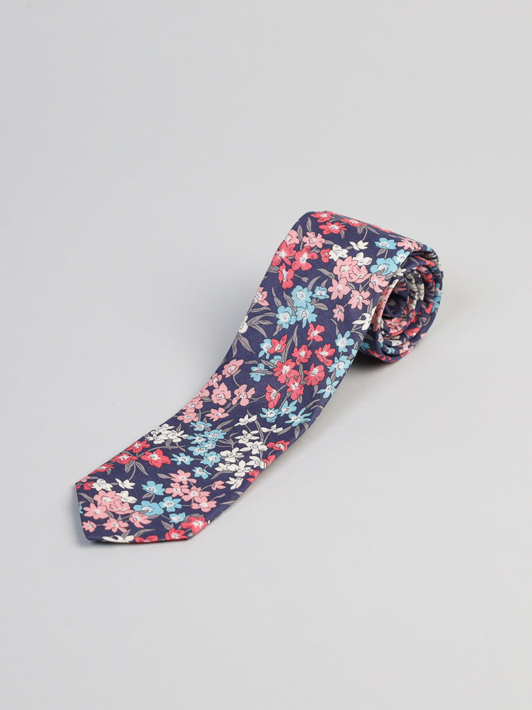 The ID Liberty of London Tana Lawn™ Sea Bloom Cotton Tie by ID Menswear, featuring red, pink, blue, and white flowers on a dark blue background, is elegantly displayed against a light gray backdrop. Handmade in Brooklyn from Tana Lawn cotton, it exudes charm.