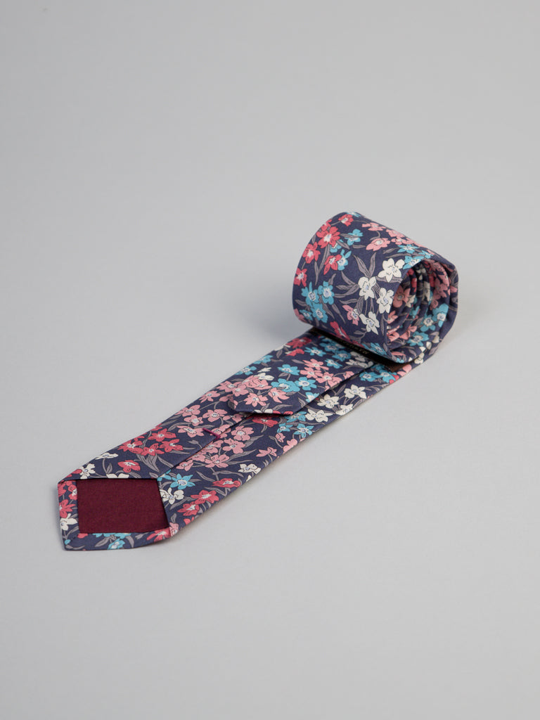 The Liberty of London Tana Lawn™ Sea Bloom Tie by ID Menswear, featuring a vibrant red, blue, and white floral pattern on soft cotton, is rolled up on a light gray surface with a dark background.