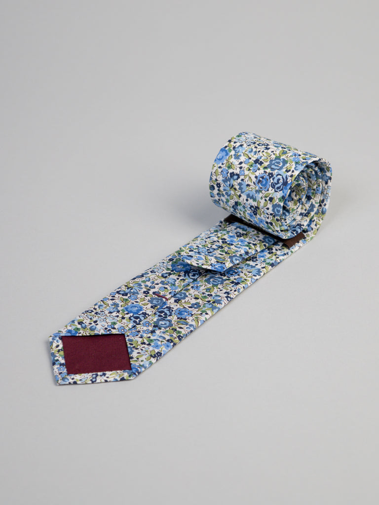The Liberty of London Tana Lawn™ Emma Tie by ID Menswear, handmade in Brooklyn, features a blue and green floral pattern on white cotton with a maroon underside tip, shown rolled up on a light gray surface.