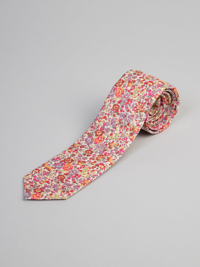 The ID Menswear Liberty of London Tana Lawn™ Georgina Tie features a colorful floral pattern in pink, red, orange, and yellow on a light background. Handmade in Brooklyn, it is displayed rolled up on a plain gray surface.
