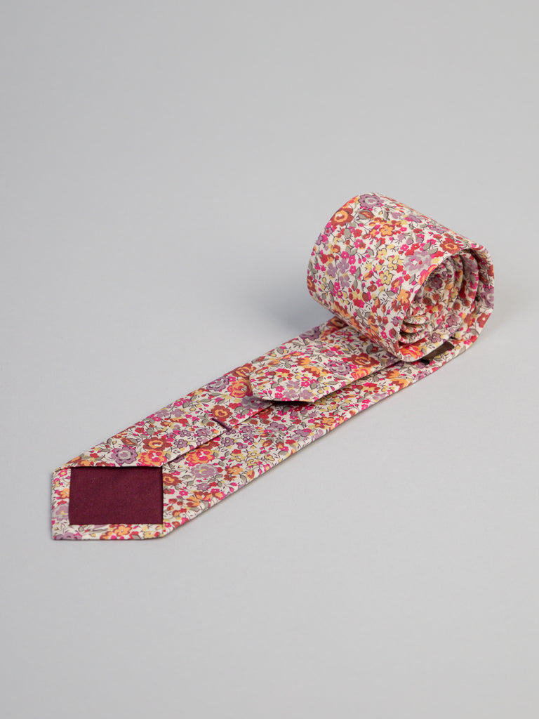 The Liberty of London Tana Lawn™ Georgina Tie by ID Menswear features a colorful floral print in pink, red, orange, and yellow on a light background, handmade in Brooklyn and displayed rolled up on a plain gray surface.