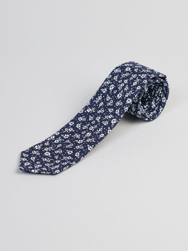 The Ditsy Cotton Tie by ID Menswear, handmade in Brooklyn, features a rolled-up navy blue cotton fabric with a white floral pattern and is displayed on a light gray background.